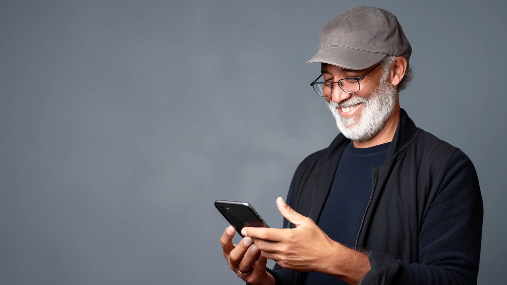 Older man with a grey cap and glasses smiling while using a smartphone against a grey background